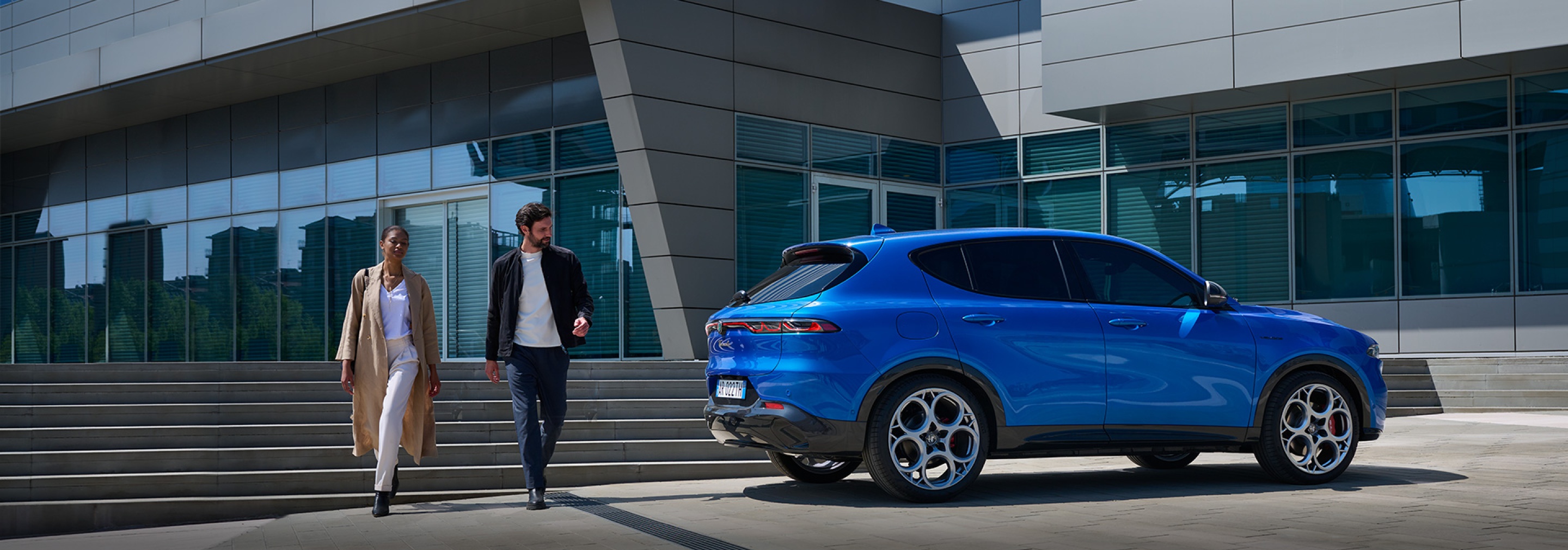 Kobieta i meżczyzna na tle schodó do biurowca, obok błędkitne auto z marek oferowanych przez grupę Stellantis