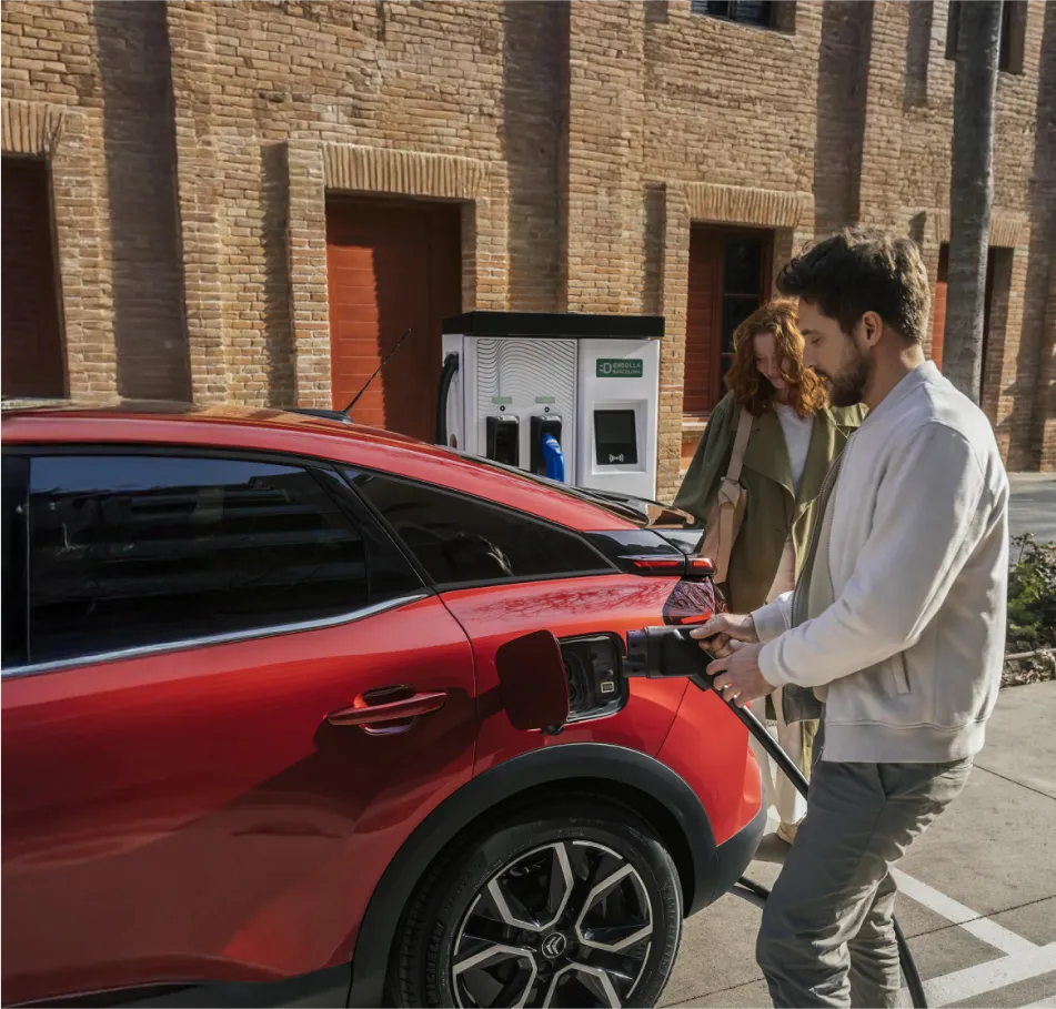 Męzczyzna ładuje czerwone auto elektryczne, za nim stoi kobieta